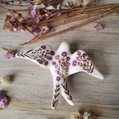 Pássaro de cerâmica branca com pintura floral roxa e castanha sobre madeira clara com flores secas