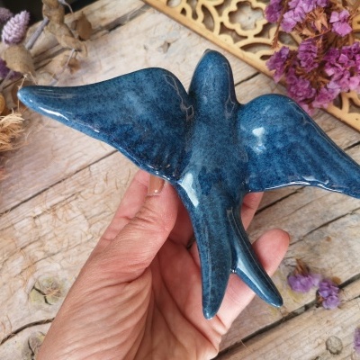 Escultura de pássaro azul em cerâmica segurada à mão sobre madeira e flores secas