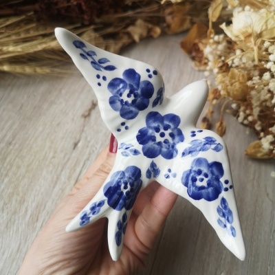 Escultura de pássaro em cerâmica branca com flores azuis, segurada por mão, sobre madeira clara.