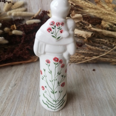 Figura de cerâmica branca com flores vermelhas e verdes, pessoa a segurar criança, sobre madeira clara com plantas secas