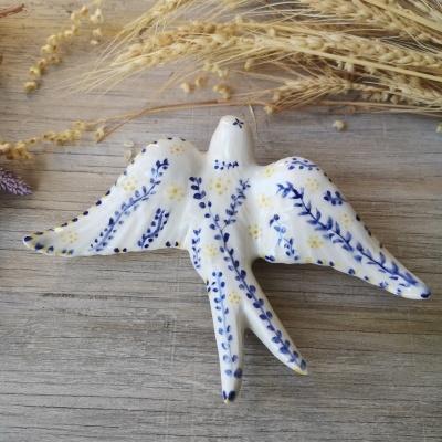 Figura decorativa de pássaro em cerâmica branca com pintura azul e amarela sobre superfície de madeira.
