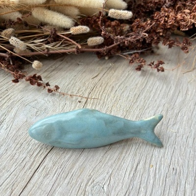 Elemento decorativo cerâmico azul esverdeado em forma de peixe sobre madeira clara com plantas secas ao fundo