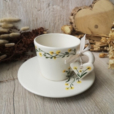 Conjunto de chávena cerâmica branca decorada com flores amarelas e pires junto a coração pendurado e fundo natural