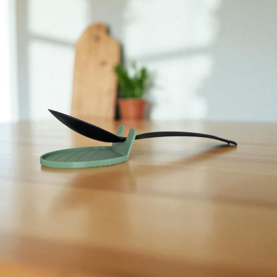 Espátula de cozinha preta com suporte verde em forma de folha sobre mesa de madeira.