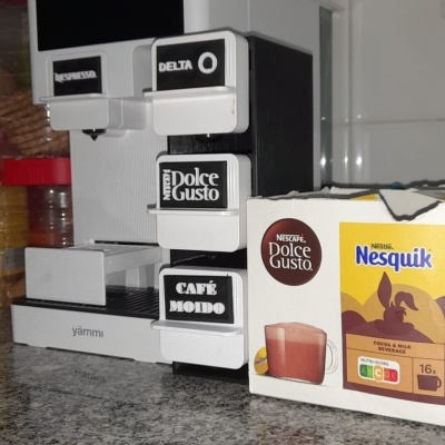 Máquina de café branca e preta com botões de diferentes marcas e caixa de cápsulas Nesquik na bancada.