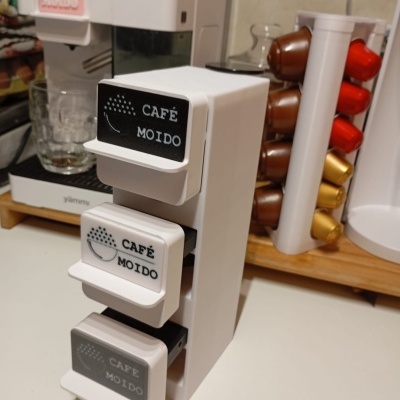 Suporte com caixas para café moído ao lado de máquina de café e cápsulas coloridas numa cozinha