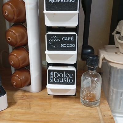 Suporte de cápsulas de café com etiquetas e cápsulas castanhas ao lado de garrafa pequena em superfície de madeira