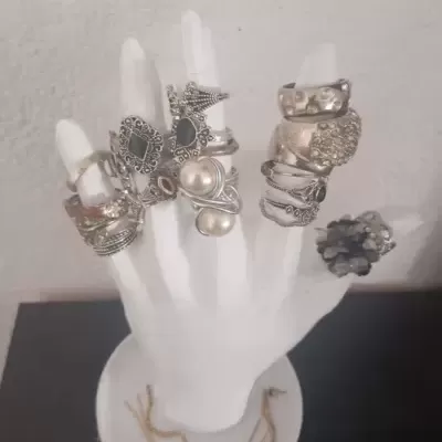 Mão expositora branca com anéis variados e pulseiras na base.