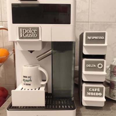 Máquina de café branca NESCAFÉ Dolce Gusto com cápsulas Nespresso, Delta e Café Moído numa bancada de cozinha.