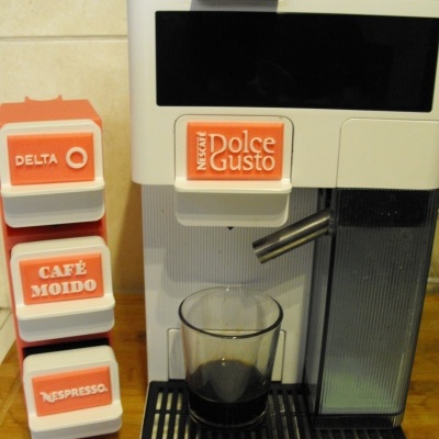 Máquina de café branca com copo de café e cápsulas com etiquetas laranja e branco sobre bancada