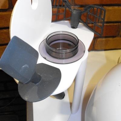 Acessório branco e cinzento para eletrodoméstico com recipiente circular e grelha, sobre superfície branca e parede de tijolo.