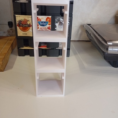 Suporte branco de plástico com três prateleiras numa bancada de cozinha com saquetas de chá ao fundo.