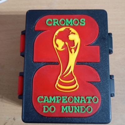 Caixa preta e vermelha com texto CROMOS 82 CAMPEONATO DO MUNDO e relevo da Taça do Mundo