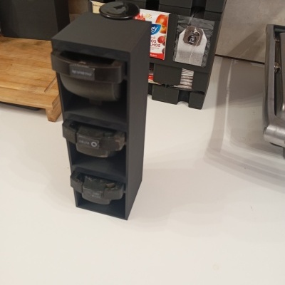 Suporte preto vertical com cápsulas de café Nespresso, numa bancada branca, perto de um tabuleiro de madeira e pacotes de chá