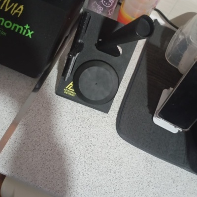 Suporte de mesa preto com vários compartimentos, copos de plástico coloridos e objetos eletrónicos numa superfície cinzenta.