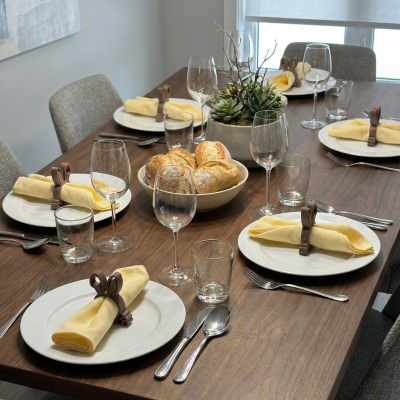 Mesa de jantar posta com pratos, guardanapos amarelos em argolas de coelho, talheres, copos de água e vinho, tigela de pães e centro de mesa com plantas suculentas.
