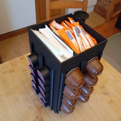 Organizador preto para cápsulas de café e açúcar com cápsulas castanhas e roxas e pacotes laranja com texto azul e branco numa mesa de madeira