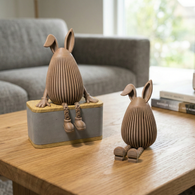 Duas figuras decorativas de coelhos castanhos sentados numa mesa de madeira numa sala com sofá cinzento.