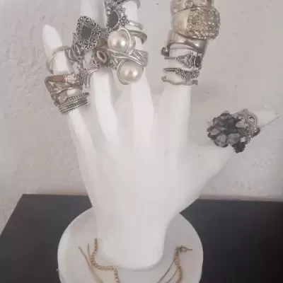 Mão exibidora branca com vários anéis em diferentes estilos sobre base circular branca com colares dourados