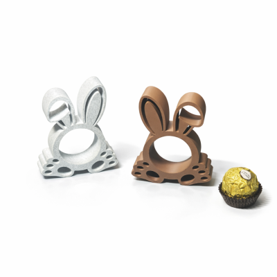 Duas argolas de guardanapo em forma de coelho, uma cinzenta e outra castanha, ao lado de um bombom dourado.