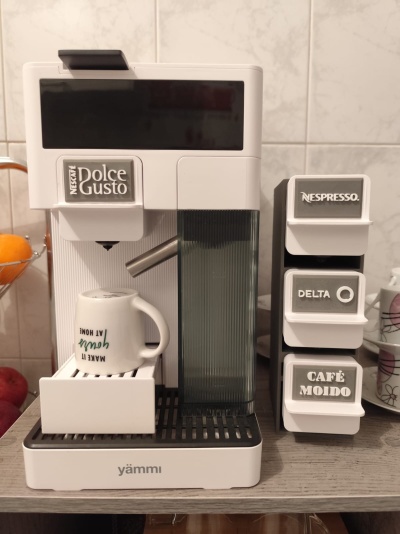 Máquina de café branca NESCAFÉ Dolce Gusto com cápsulas Nespresso, Delta e Café Moído numa bancada de cozinha.