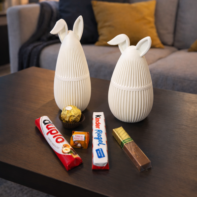 Dois recipientes de coelho brancos estriados com chocolates variados numa mesa de madeira escura.