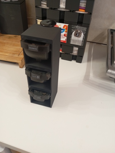 Suporte preto vertical com cápsulas de café Nespresso, numa bancada branca, perto de um tabuleiro de madeira e pacotes de chá