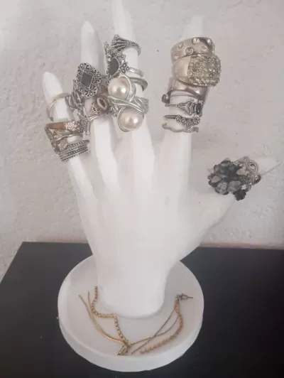 Mão exibidora branca com vários anéis em diferentes estilos sobre base circular branca com colares dourados