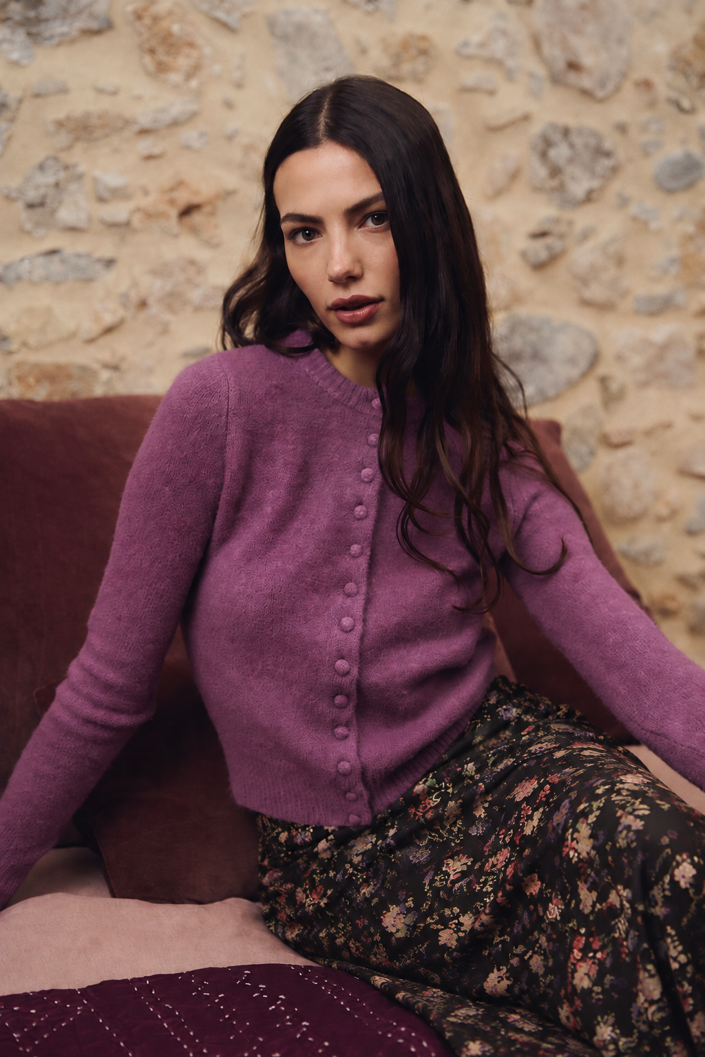 CASACO BONIFÁCIA MALVA Mulher com casaco roxo e saia floral sentada num sofá com almofadas castanhas.