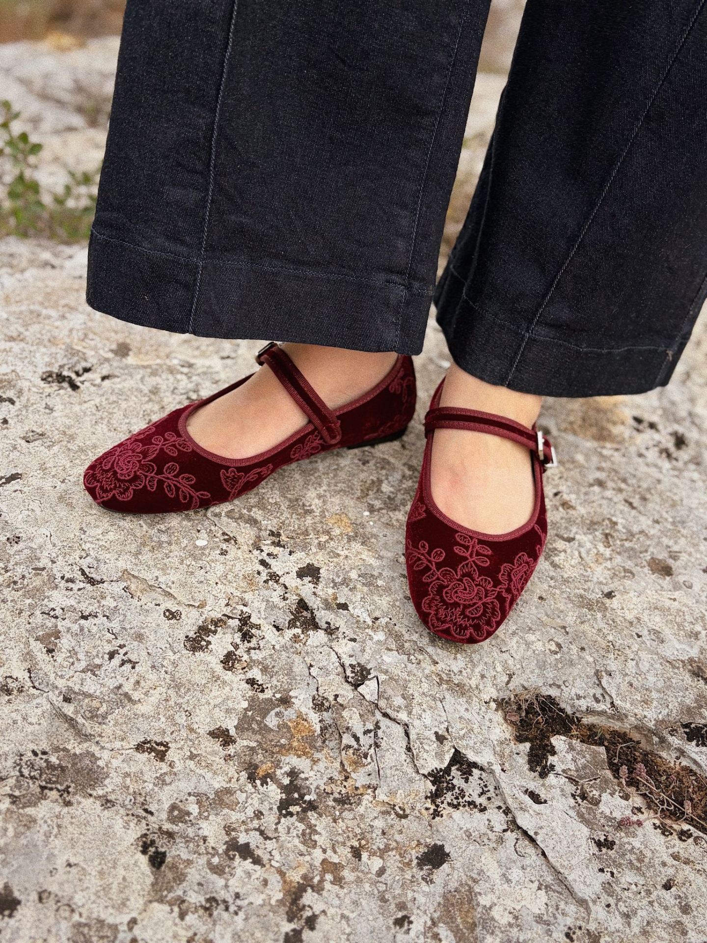 MARY-JANE VELVET BORDEAUX Sapatos Mary Jane vermelhos em veludo bordado usados com calças pretas sobre chão de pedra.
