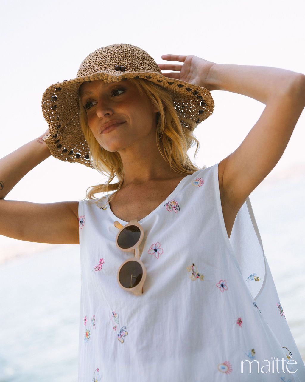 TÚNICA AIDÉ Mulher com vestido branco floral, óculos de sol bege, chapéu de palha castanho claro ao ar livre de frente para o mar.