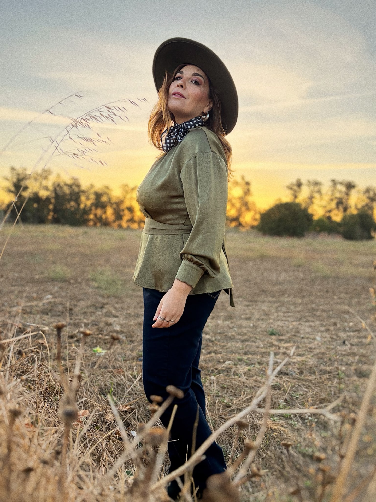 BLUSA BINA VERDE KHAKI Mulher jovem com camisa verde, calças escuras, lenço preto com padrão branco e chapéu castanho em campo ao pôr do sol