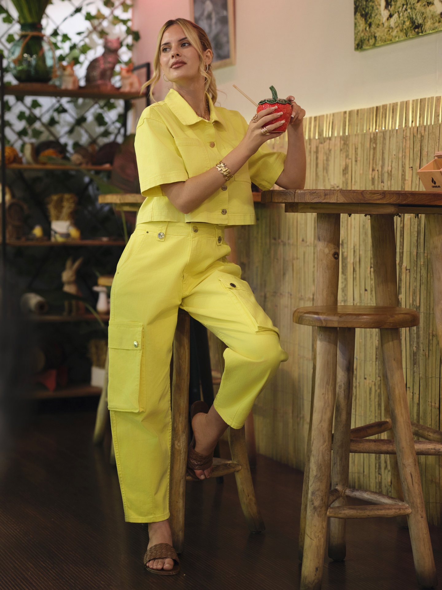 CALÇAS SOLINE Mulher vestindo conjunto amarelo com calças cargo e camisa, sentada em banco de madeira, segurando caneca vermelha.