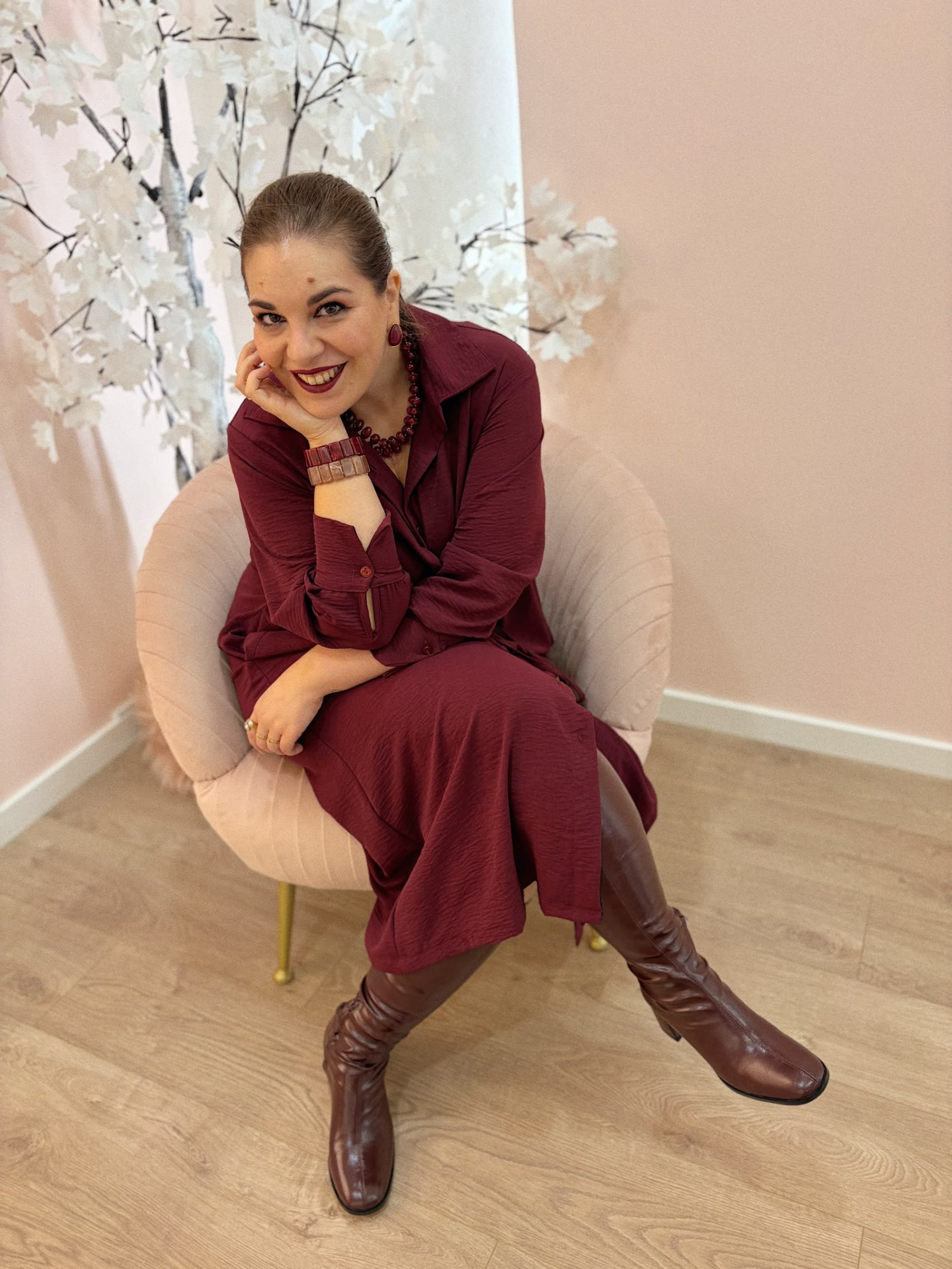 VESTIDO BRICE BORDEAUX Mulher sentada em poltrona rosa usando vestido vinho e botas castanhas.
