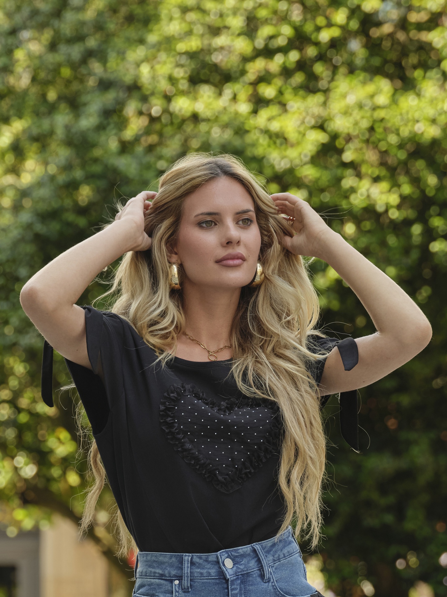 T-SHIRT SÓNIA Mulher loira de camisola preta com coração e calças de ganga azuis ao ar livre