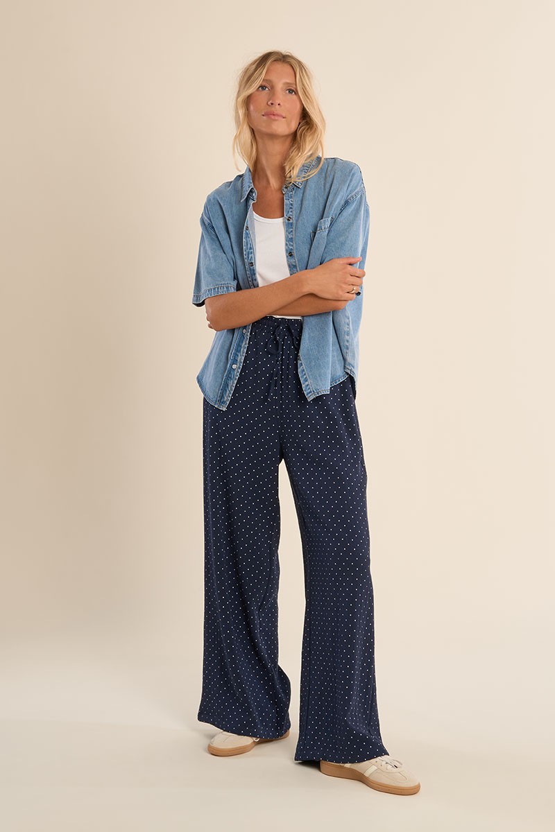 CALÇAS YOLANDA Mulher com camisa de ganga azul clara, t-shirt branca, calças largas azuis com pontos brancos e sapatilhas claras
