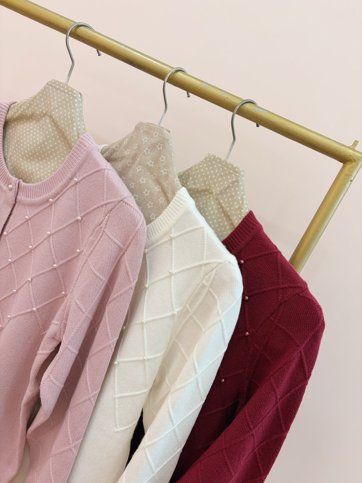 CASACO ARINDA Três cardigans de malha rosa claro, branco e vermelho pendurados em cabides revestidos de tecido bege estampado com estrelas.