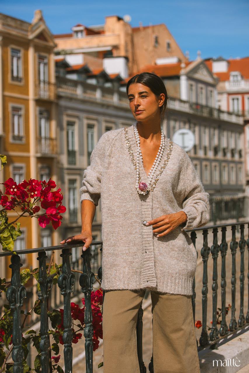 CASACO CHLOE Mulher com cardigã cinza e colar de pérolas em varanda urbana com flores vermelhas