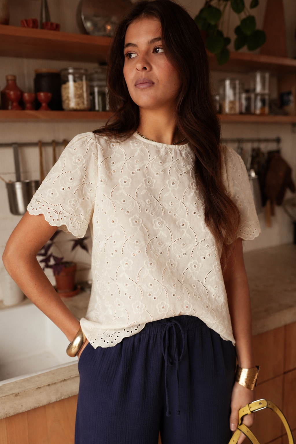 BLUSA VILMA Blusa branca bordada com calças azuis e acessórios dourados numa cozinha.