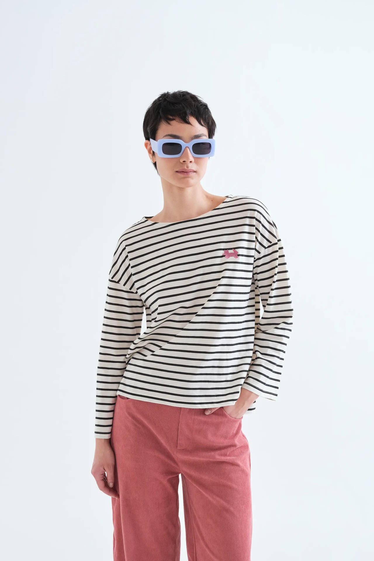 CAMISOLA RAYAS NEGRAS Pessoa com óculos de sol azuis, camisa às riscas pretas e brancas com cão rosa e calças de veludo cotelê rosa