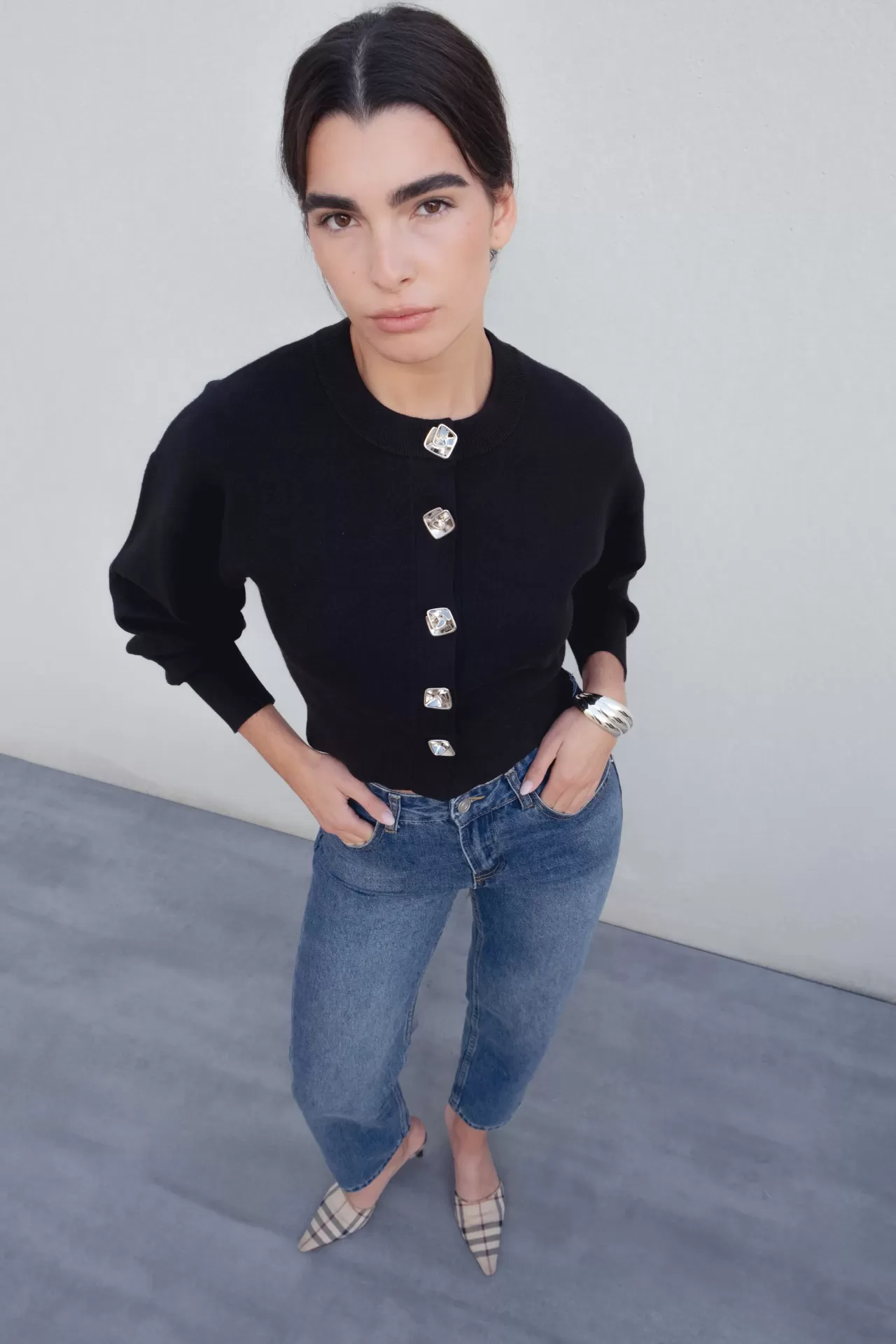 CASACO RUNA Mulher com camisola preta abotoada, jeans azuis e sapatos mule xadrez em fundo cinza