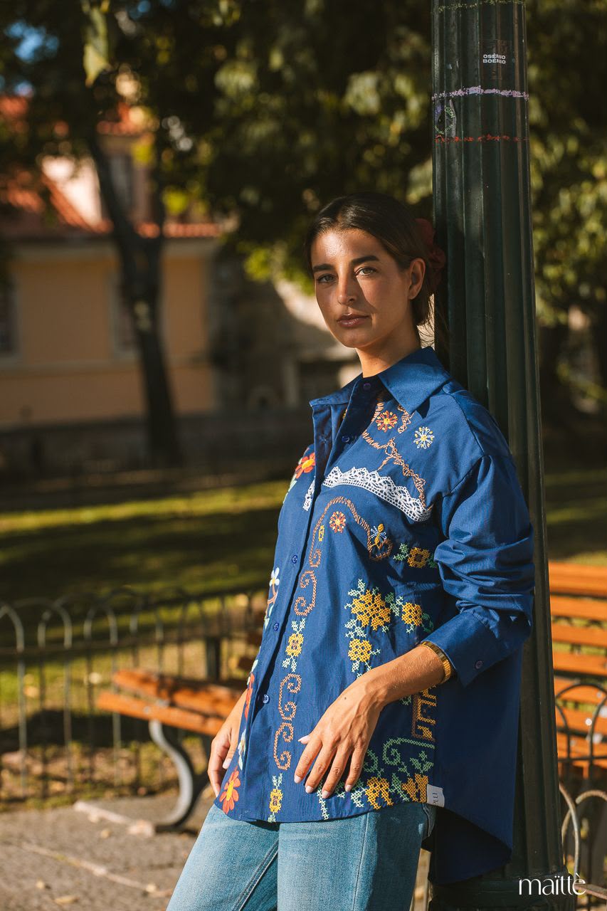 CAMISA CLAIRE Camisa azul bordada com padrões florais e geométricos coloridos, com renda branca, usada ao ar livre