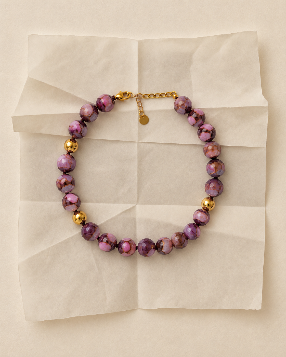 COLAR APOLÓNIA ROSA Colar de contas facetadas lilás, rosa, castanho e dourado sobre papel bege.