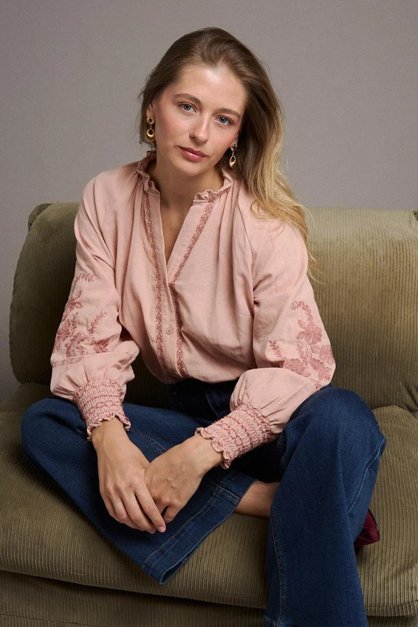 BLUSA AVA Mulher sentada num sofá verde-claro a usar blusa rosa clara bordada e jeans azul escuro