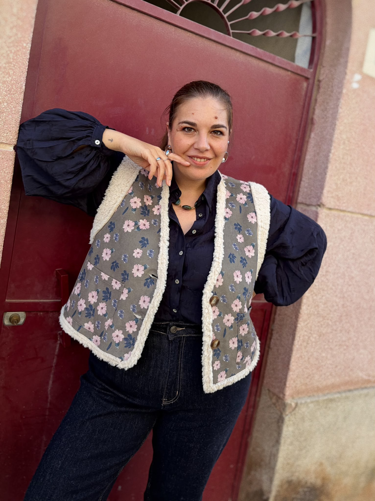 BLUSA CHARLENE Mulher com colete floral cinzento com detalhes felpudos e camisa preta em frente a porta vermelha