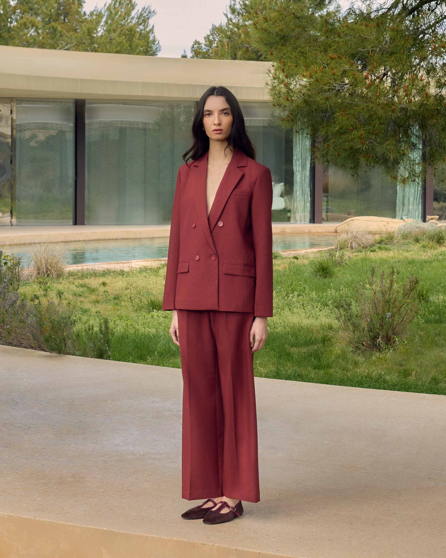 BLAZER NOIX TERRACOTA Mulher com fato castanho avermelhado e sapatos planos castanhos em camurça, em ambiente exterior com vegetação e piscina.