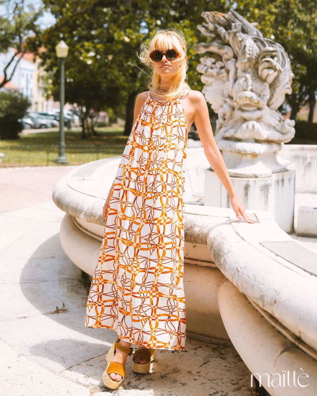 VESTIDO AGNA Mulher usando vestido longo branco com padrão laranja, sandálias camel e óculos escuros, ao lado de fonte de pedra