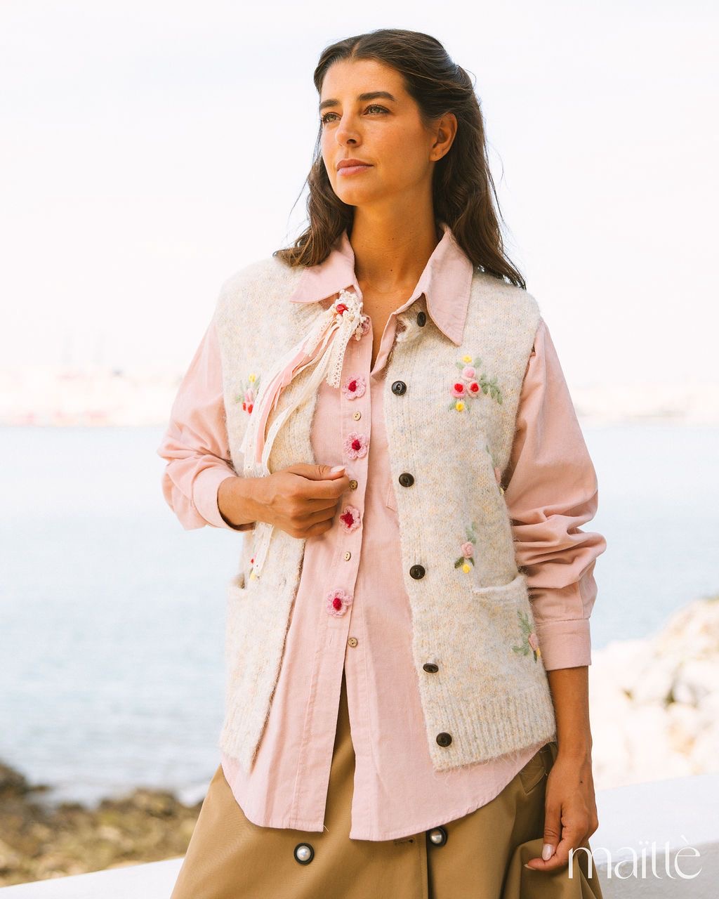 CAMISA CATALEIA Colete bege com bordados florais usado sobre camisa rosa e calças castanhas em ambiente à beira-mar.
