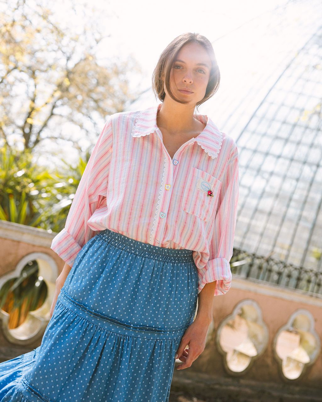 CAMISA STELINA Mulher com blusa riscada rosa e branca com gola e renda, saia azul de bolinhas brancas ao ar livre.