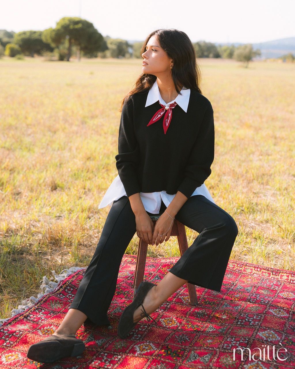 CONJUNTO ASSUNTA Mulher sentada na relva com roupa preta e branca e lenço vermelho, sobre tapete vermelho com padrão.
